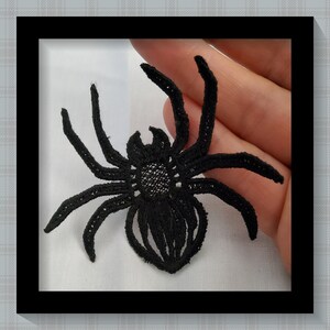 Könnte beinhalten: Schwarze Spitzenapplikation einer Spinne mit acht Beinen und einem detaillierten Körper. Die Spinne befindet sich vor einem weißen Hintergrund in einem schwarzen Rahmen. Das filigrane Design der Spinne ist sichtbar.