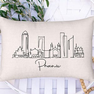 May include: A rectangular, cream-colored pillow featuring a black line drawing of the Phoenix, Arizona skyline. The city's name is written in cursive below the skyline. The pillow is placed on a white, circular decorative tray.