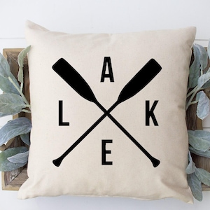 May include: A beige square pillow with the word "LAKE" in black block letters, with crossed black oars. The pillow is displayed on a wooden tray with decorative greenery and wooden beads.