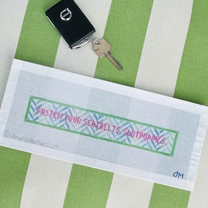 May include: A white needlepoint canvas with the text "FASTEN YOUR SEATBELTS SHUTUPPIES" in pink and blue, framed in green. A black car key and key are on a green and white striped surface.