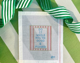 May include: A needlepoint canvas with the text "DO IT WHILE YOU'RE GOOD & PISSED" in blue, red, and white. A green and white striped ribbon is draped over the canvas, which is on a green and white striped background.