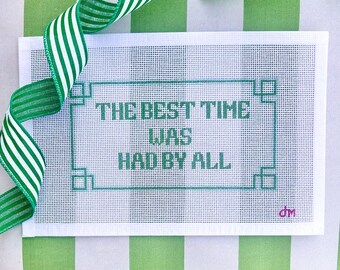 Op de afbeelding: Een wit borduurcanvas met de tekst "THE BEST TIME WAS HAD BY ALL" in teal. De tekst is omlijst door een teal rand. Een groen-wit gestreept lint is over het canvas gedrapeerd.