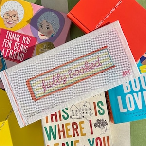 May include: A white needlepoint bookmark with the phrase "fully booked" in pink and yellow, framed by a red border. It's placed on a pile of books, including one with the text "Thank You For Being A Friend".