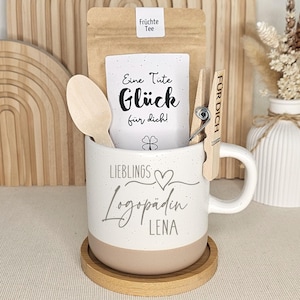 Könnte beinhalten: Eine weiße Keramiktasse mit beigem Boden, gefüllt mit einem Teebeutel, einem Holzlöffel und einer Wäscheklammer. Auf der Tasse steht "LIEBLINGS Logopädin LENA". Unter der Tasse befindet sich ein Untersetzer aus Holz.