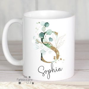 Könnte beinhalten: Weiße Keramiktasse mit einem goldenen Glitzerbuchstaben "S", umgeben von grünen Eukalyptusblättern und goldenen Akzenten. Der Name "Sophia" ist in schwarzer Kursivschrift unter dem Buchstaben geschrieben.