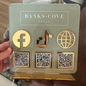 May include: A decorative sign for Banks + Cove Pet Co. features a light green background with gold accents. The sign includes a Facebook logo, a TikTok logo, and a globe icon, along with three QR codes. The text "BANKS + COVE PET CO." is displayed at the top.