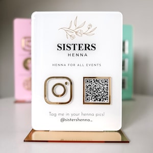 Op de afbeelding: Een wit acryl bord met gouden accenten met de tekst "Sisters Henna" en "Henna voor alle evenementen". Het bord bevat ook een Instagram-logo en een QR-code. De tekst "Tag me in your henna pics! @sistershenna_" staat onderaan het bord.