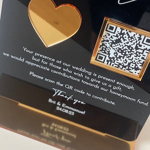May include: Black and gold wedding invitation with a heart and a QR code. The text reads: "Your presence at our wedding is present enough, but for those who wish to give us a gift, we would appreciate contributions towards our honeymoon fund. Please scan the QR code to contribute. Thank you Brii & Emmanuel 24.08.23"