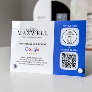 May include: A clear acrylic sign with a white and blue design. The sign features the text "by Victoria Maxwell" and prompts for a Google review. It includes options to tap a phone via NFC or scan a QR code.