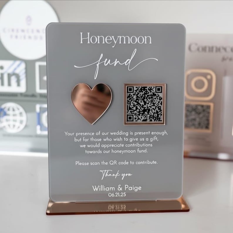 May include: A clear acrylic sign with a rose gold base and accents. The sign reads "Honeymoon fund" with a heart and QR code. Text below asks guests to scan the code to contribute. Names and a date are printed at the bottom.