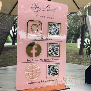 May include: Pink acrylic sign with gold lettering and icons for Bay Laurel Wedding + Events. The sign includes social media icons for Instagram, Facebook, and a website, along with QR codes for each. The text "by Victoria Maxwell" is at the bottom of the sign.