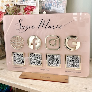 May include: A pink acrylic sign with the name "Suzie Marie" written in black cursive. The sign has gold icons for Facebook, Instagram, and Google, as well as a globe icon. Below the icons are four QR codes. The sign also includes the text "www.suziemarie.co.uk", "Let's get social", and "Please leave a review".