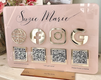 May include: A pink acrylic sign with the name "Suzie Marie" written in black cursive. The sign has gold icons for Facebook, Instagram, and Google, as well as a globe icon. Below the icons are four QR codes. The sign also includes the text "www.suziemarie.co.uk", "Let's get social", and "Please leave a review".