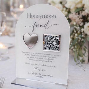 Può includere: Segno di matrimonio in acrilico trasparente con testo "Honeymoon fund", un ritaglio a forma di cuore e un codice QR. Il cartello include il testo che chiede agli ospiti di contribuire al fondo luna di miele. Sono stampati anche i nomi Conchata & Lawrence e la data 08.23.26.