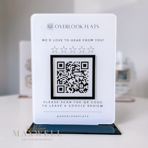 May include: White sign with black text that reads "OVERLOOK FLATS" and "WE'D LOVE TO HEAR FROM YOU!" with five black stars. A black and white QR code is below the text. The text below the QR code reads "PLEASE SCAN THE QR CODE TO LEAVE A GOOGLE REVIEW" and "@OVERLOOKFLATS".