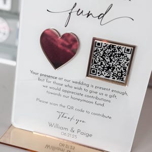May include: A white card with the text "Honeymoon fund" in black cursive font. A pink heart and a gold square with a QR code are on the card. The text "Your presence at our wedding is present enough, but for those who wish to give us a gift, we would appreciate contributions towards our honeymoon fund. Please scan the QR code to contribute. Thank you William & Paige 06.21.25" is printed on the card.