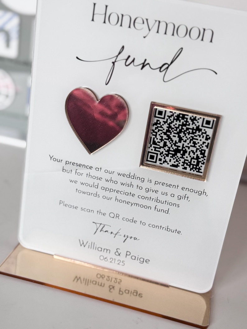 May include: A white card with the text "Honeymoon fund" in black cursive font. A pink heart and a gold square with a QR code are on the card. The text "Your presence at our wedding is present enough, but for those who wish to give us a gift, we would appreciate contributions towards our honeymoon fund. Please scan the QR code to contribute. Thank you William & Paige 06.21.25" is printed on the card.