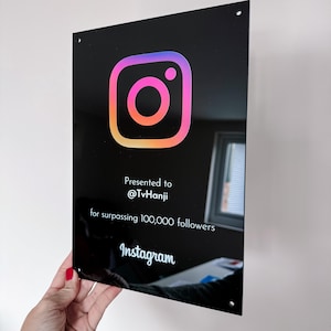 May include: Black plaque with the Instagram logo in rainbow colors. The text "Presented to @TvHanji for surpassing 100,000 followers" is written in white. The word "Instagram" is written in white at the bottom.