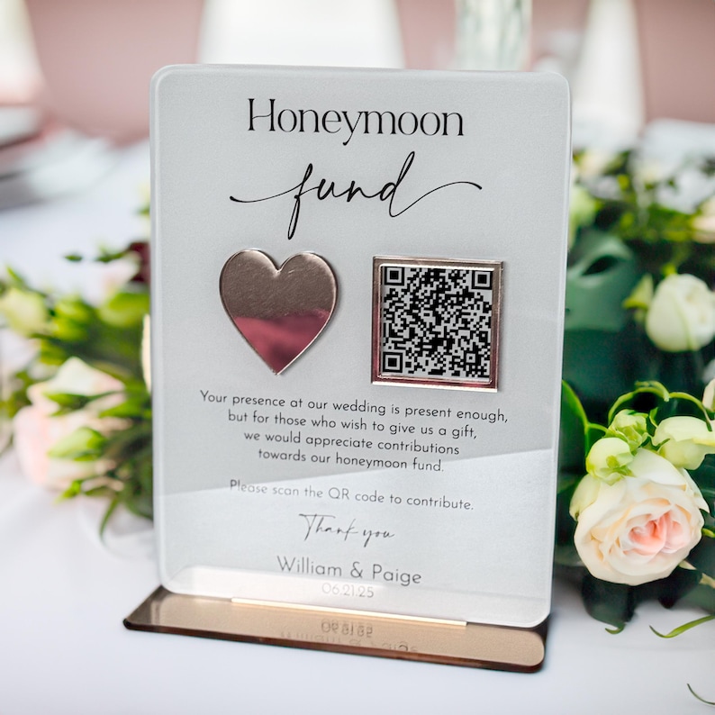 May include: A white acrylic sign with the words "Honeymoon Fund" in black text. The sign has a silver heart and a QR code. The text below the QR code reads "Your presence at our wedding is present enough, but for those who wish to give us a gift, we would appreciate contributions towards our honeymoon fund. Please scan the QR code to contribute. Thank you William & Paige 0621/25"