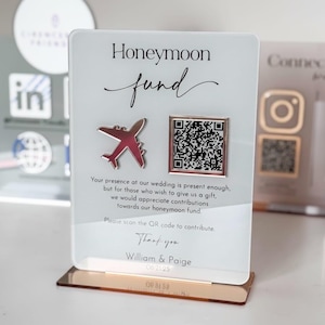 Puede incluir: Un letrero de acrílico transparente con una base de oro rosa, con las palabras "Honeymoon fund" y un gráfico de avión rojo. Incluye un código QR y texto que invita a contribuciones para una luna de miel.