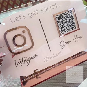 May include: A pink and gold acrylic sign with the text "Let's get social..." and a QR code with the text "Scan Here". The sign also features an Instagram logo and the text "@by.kndl" and "Instagram".