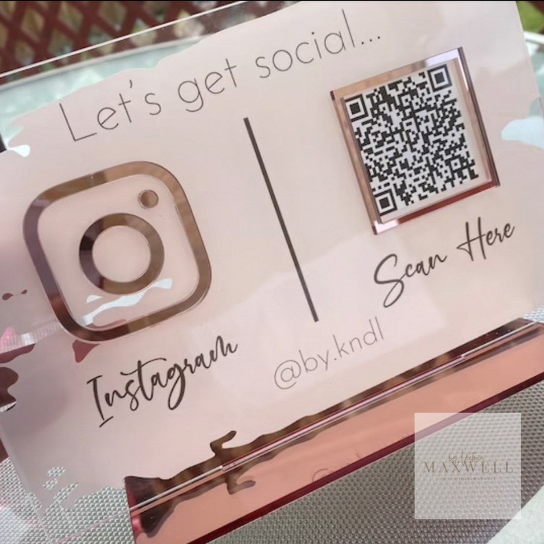 May include: A pink and gold acrylic sign with the text "Let's get social..." and a QR code with the text "Scan Here". The sign also features an Instagram logo and the text "@by.kndl" and "Instagram".