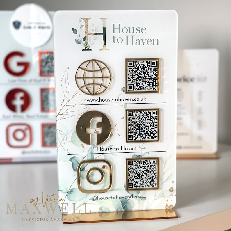Op de afbeelding: Een witte acryl displaystandaard met gouden accenten met de tekst "House to Haven" en social media-iconen, waaronder een wereldbol, Facebook en Instagram. De standaard bevat ook QR-codes voor elk social media-platform. De tekst "by Victoria Maxwell" en het websiteadres "www.housetohaven.co.uk" zijn ook op de standaard te vinden.