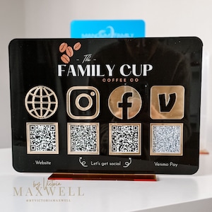 Może przedstawiać: Czarny znak z złotymi akcentami z tekstem "The Family Cup Coffee Co." z ikoną globusa, Instagrama, Facebooka i Venmo. Pod ikonami znajdują się cztery kody QR oznaczone jako "Website", "Let's get social" i "Venmo Pay". Znak znajduje się na brązowym stojaku z tekstem "by Victoria MAXWELL @BYVICTORIAMAXWELL" na dole.