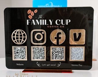May include: Black sign with gold accents featuring the text "The Family Cup Coffee Co." with a globe, Instagram, Facebook, and Venmo icons. Below the icons are four QR codes labeled "Website", "Let's get social", and "Venmo Pay". The sign is on a brown stand with the text "by Victoria MAXWELL @BYVICTORIAMAXWELL" at the bottom.