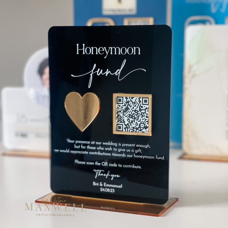 May include: Black acrylic sign with gold accents, reading "Honeymoon fund". A gold heart and a QR code are displayed. The text reads "Your presence at our wedding is present enough, but for those who wish to give us a gift, we would appreciate contributions towards our honeymoon fund. Please scan the QR code to contribute. Thank you Brii & Emmanuel 24.08.23"