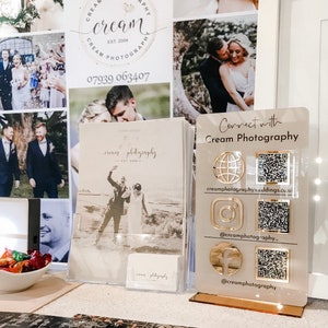 Op de afbeelding: Een wit bord met gouden accenten toont de tekst "Connect with Cream Photography" en bevat een websiteadres, social media-handvatten en QR-codes voor elk platform. Het bord staat op een tafel met een verscheidenheid aan foto's en andere promotiematerialen.