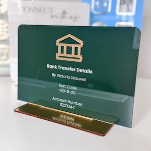 May include: A dark green acrylic bank transfer details sign with gold accents. The sign features a gold bank building icon and the text "Bank Transfer Details" and "By Victoria Maxwell". The sign also displays a sort code and account number.
