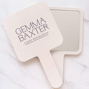 Può includere: Uno specchio a mano color crema con uno specchio rettangolare e un manico. Il testo "GEMMA BAXTER" è stampato in viola scuro, insieme a "ELEGANT WEDDING MAKEUP & BRIDAL SKIN SPECIALIST".