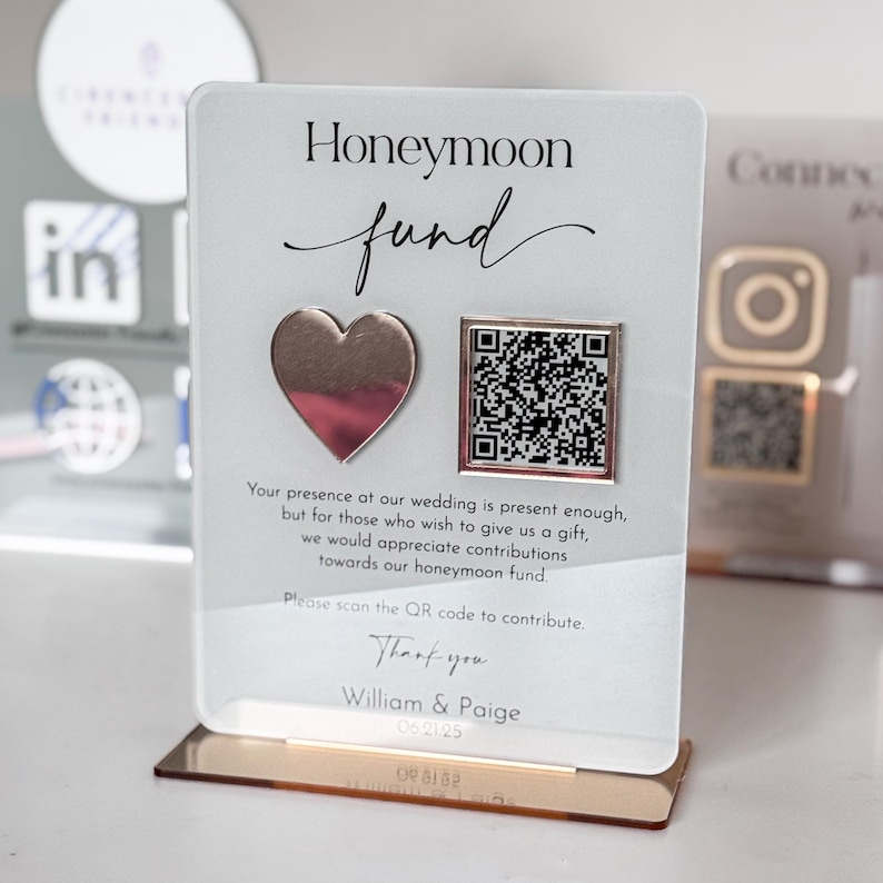 May include: A white acrylic sign with the text "Honeymoon Fund" in black script font. The sign has a pink and silver heart and a black and white QR code. The text "Your presence at our wedding is present enough, but for those who wish to give us a gift, we would appreciate contributions towards our honeymoon fund. Please scan the QR code to contribute. Thank you William & Paige 0621/25" is printed on the sign.