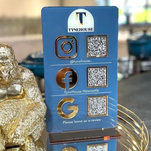 Op de afbeelding: Een blauw acryl bord met gouden letters en pictogrammen. Het bord heeft de tekst "TyneHouse" en bevat sociale media pictogrammen voor Instagram, Facebook en Google. Het bord bevat ook QR-codes voor elk sociaal media platform. De tekst "Laat ons een beoordeling achter" staat onderaan het bord.