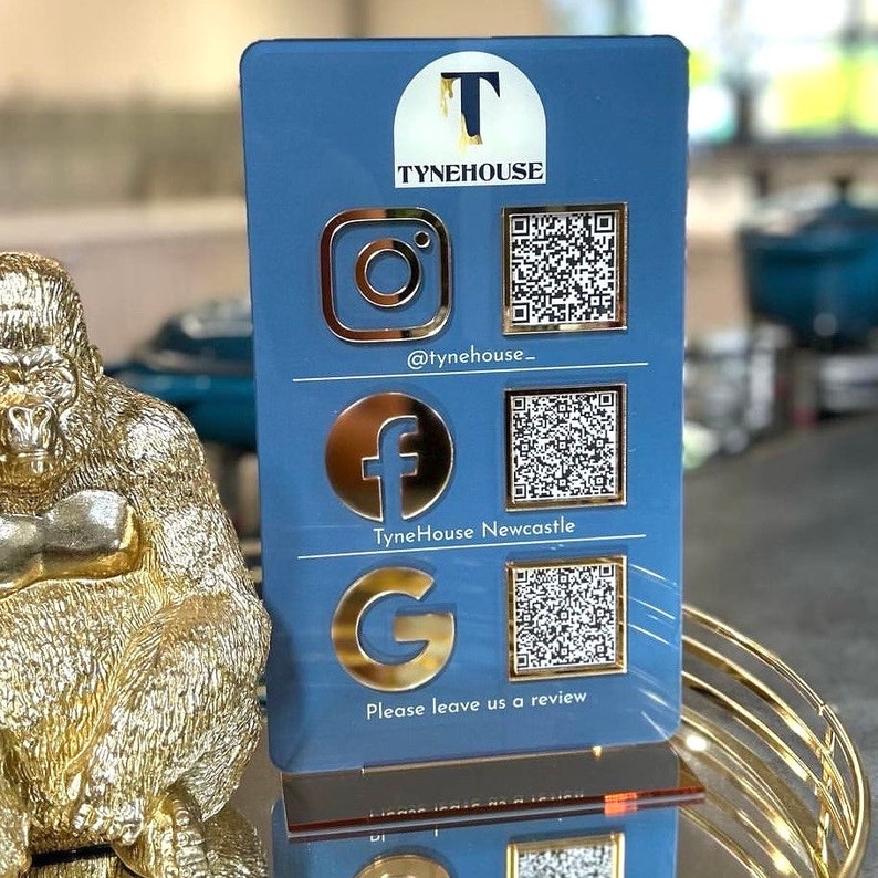 Op de afbeelding: Een blauw acryl bord met gouden letters en pictogrammen. Het bord heeft de tekst "TyneHouse" en bevat sociale media pictogrammen voor Instagram, Facebook en Google. Het bord bevat ook QR-codes voor elk sociaal media platform. De tekst "Laat ons een beoordeling achter" staat onderaan het bord.