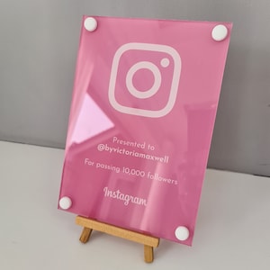 May include: Pink acrylic plaque with a white Instagram logo, the text "Presented to @byvictoriamaxwell For passing 10,000 followers Instagram" and white dots on the corners. The plaque is on a small wooden easel.