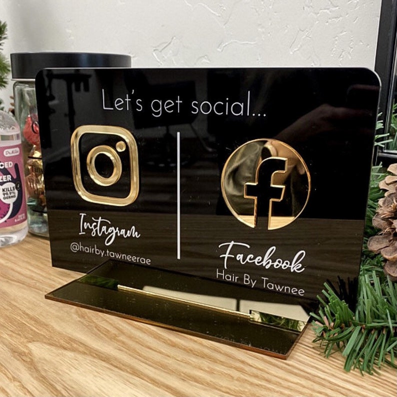 May include: Black acrylic sign with gold accents, featuring Instagram and Facebook logos and handles. The sign reads "Let's get social..." and includes the text "Instagram @hairby.tawnneerae" and "Facebook Hair By Tawnee".