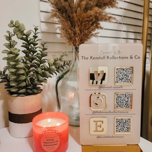 Op de afbeelding: Een roze kaars met de tekst "Pink Prosecco Frosting" op het etiket. Een houten bord met de tekst "Connect With The Kendall Kollections & Co." en social media-handvatten en QR-codes.