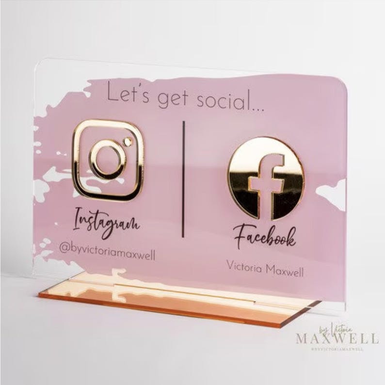 May include: A pink acrylic sign with gold accents featuring the Instagram and Facebook logos. The sign reads "Let's get social..." with the Instagram handle "@byvictoriamxwell" and the Facebook name "Victoria Maxwell".
