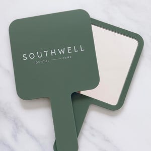 May include: A green hand mirror with the text "SOUTHWELL DENTAL CARE" in white. The mirror has a rectangular shape with rounded corners and a handle. The mirror is lying on a white and gray marble surface.