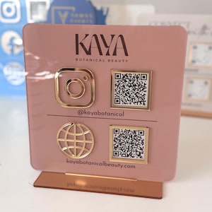 Könnte beinhalten: Ein rosa Acryl-Display mit goldfarbenen Instagram- und Globus-Symbolen sowie zwei QR-Codes. Das Display enthält den Text "KAYA BOTANICAL BEAUTY" und den Instagram-Handle "@kayabotanical". Die Website-Adresse "kayabotanicalbeauty.com" ist ebenfalls sichtbar.