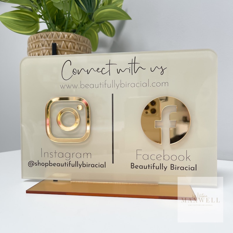 May include: A white acrylic sign with gold accents and black text. The sign says "Connect with us" and includes the website address "www.beautifullybiracial.com". The sign also features Instagram and Facebook logos with the corresponding usernames and page names.