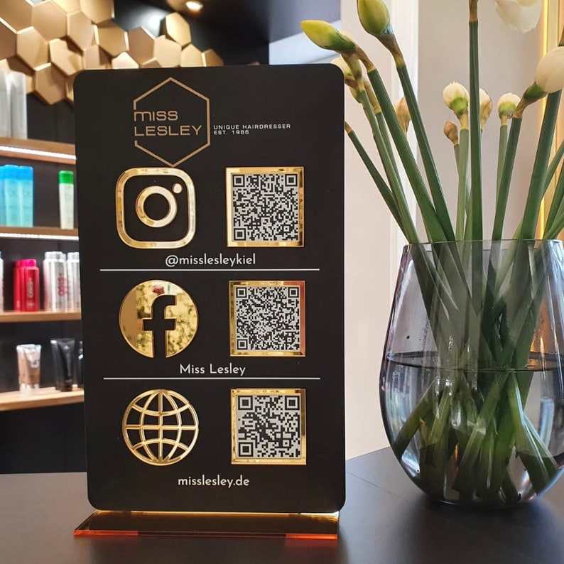 Op de afbeelding: Zwart bord met gouden letters en pictogrammen. Het bord zegt "Miss Lesley Unique Hairdresser Est. 1988". Het heeft ook pictogrammen voor Instagram, Facebook en een website, samen met QR-codes voor elk.