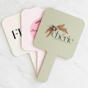 Può includere: Tre specchietti da mano. Uno è bianco sporco con la lettera "F" in nero. Il secondo è rosa chiaro con la scritta "MAKE Laura" in un cerchio rosa. Il terzo è verde salvia con la scritta "chérie AESTHETICS" e un motivo floreale.