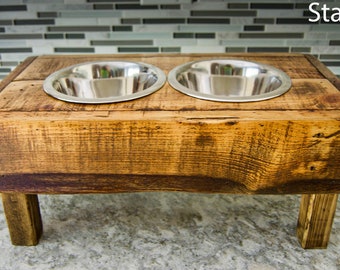 May include: A wooden dog bowl stand with two stainless steel bowls. The stand is stained a dark brown color and has a rustic look. The bowls are elevated on the stand, which is about 12 inches tall. "Stain 2" is written on the background.