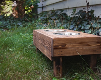 May include: A wooden dog bowl stand with two stainless steel bowls. The stand is stained a dark brown and has four legs. The stand is sitting on a bed of green grass.