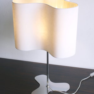 May include: A white table lamp with a wavy, organic-shaped shade and a chrome base. The lamp is turned on and the light is shining through the shade.
