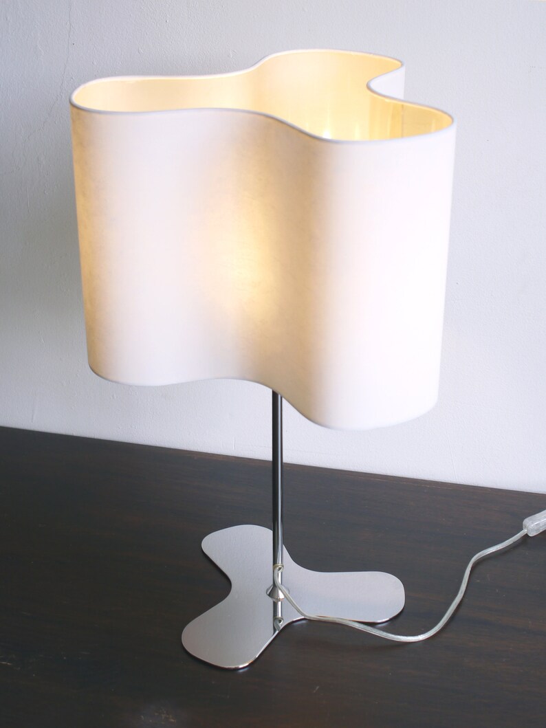 May include: A white table lamp with a wavy, organic-shaped shade and a chrome base. The lamp is turned on and the light is shining through the shade.