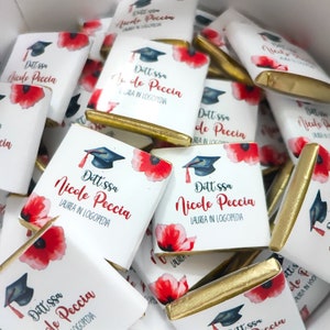 Può includere: Un mucchio di barrette di cioccolato bianco a favore con involucri in foglia d'oro. Ogni barretta presenta un fiore di papavero rosso e un cappello di laurea con il testo "Dott.ssa Nicole Peccia LAUREA IN LOGOPEDIA".